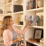 15" Classic Elephant - Side view of standing gray plush elephant being placed on a shelf in a living room by a female model image number 6